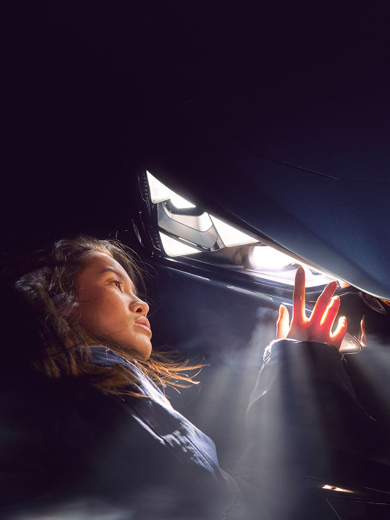 Frau in Nahaufnahme blickt auf den eingeschalteten Frontscheinwerfer eines CUPRA Tavascan, im Vordergrund das beleuchtete CUPRA Logo auf der Motorhaube